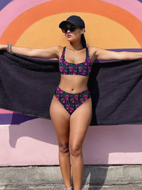 Two-piece bikini set featuring a bold pastel goth design with neon pink skulls wearing green bows, surrounded by multicolored stars on a deep black background. The set includes a plunging V-neck bikini top with wide straps and high-waisted bottoms for a flattering retro fit. Made of stretchy, soft material with a smooth black lining. This skull print swimsuit is ideal for alternative, edgy summer style with a playful punk twist.